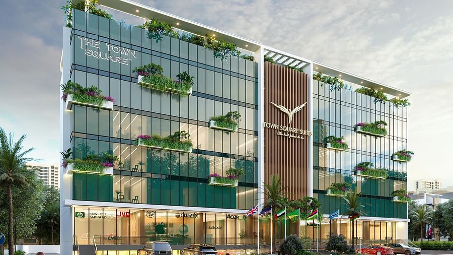 The modern multi-story exterior of Town Square Suites, featuring glass facades, green balconies, and warm dusk lighting.