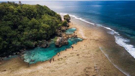 Aerial view of a sandy beach and a bright blue lagoon nestled against a forested cliff at Lamari Siargao.