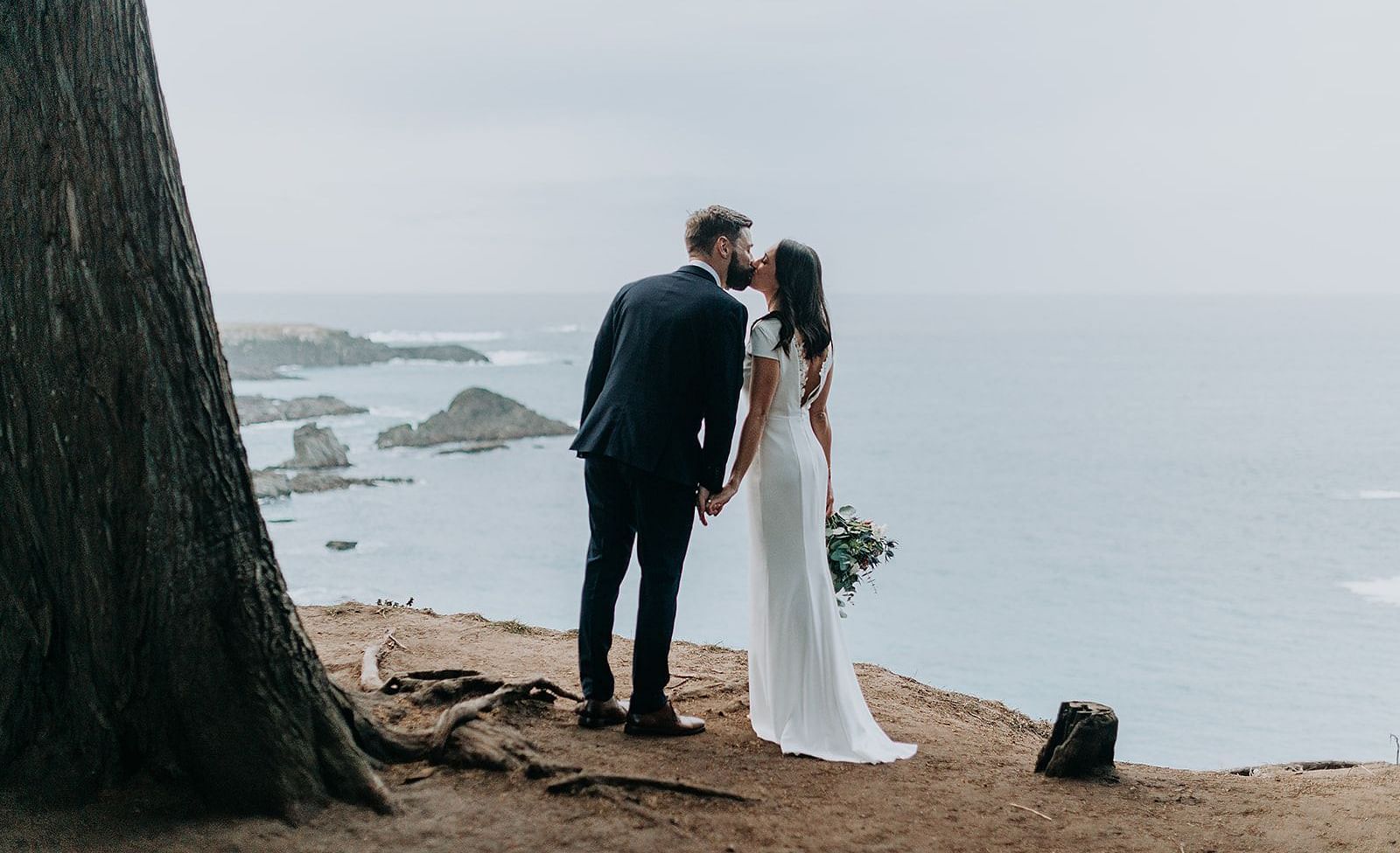 Bride and groom standing by a tree, overlooking the ocean