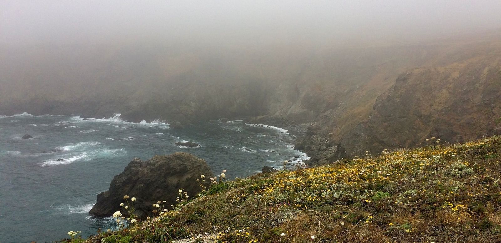 A coastal scene shows a rocky cliff descending to a body of water, with a layer of mist obscuring the horizon.