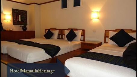 A bedroom with twin beds and a single bed, side tables, lamps, a cabinet and wall decor with the text 'Hotel Mamallaa Heritage.'