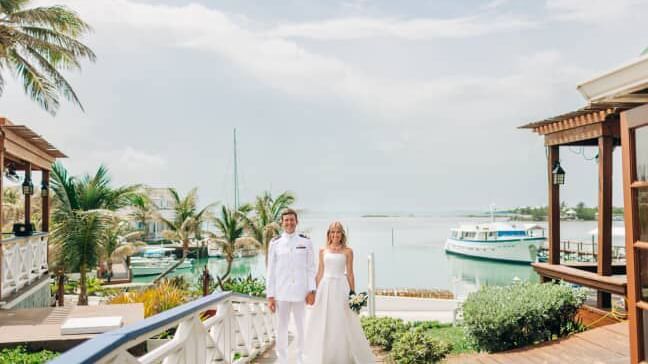 Two people in wedding attire walk on a deck lined with palm trees near boats and water at Abaco Inn.