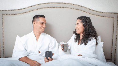 A couple in white robes enjoys hot drinks and warm smiles while relaxing in a cozy, modern room at YO1 Longevity & Health Resorts