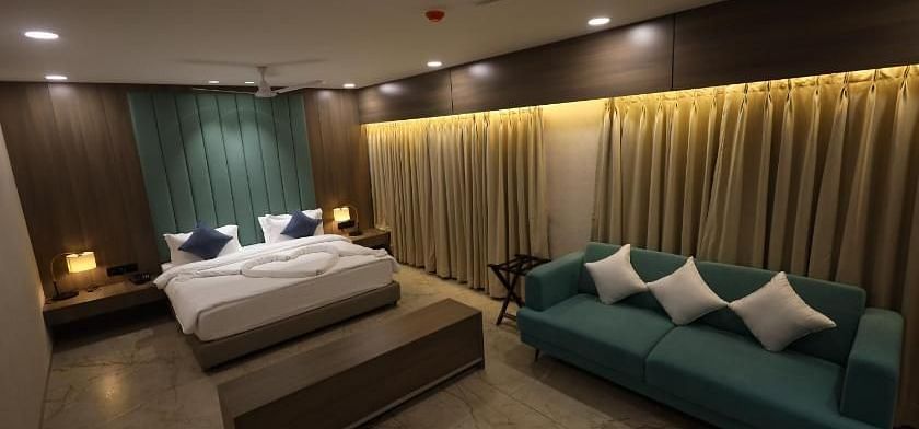 The image shows a hotel suite with a bed, a sofa with cushions, bedside tables with lamps, and curtains - Lords Eco Inn Navsari.