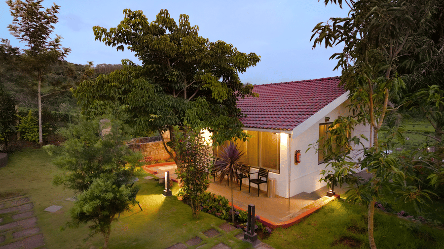 An exterior view shows a building containing the Annexe room at Ibex Resort, Kotagiri during the evening. Trees, grass, and a paved walkway surround the structure. Lights illuminate the outdoor area.