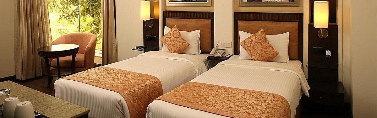 The Premium Twin Room offers twin beds with neatly arranged pillows, wooden bedside tables with lamps, a cushioned seating area with a wooden table, and large side windows for natural light at Inde Signature Tower, Gurgaon
