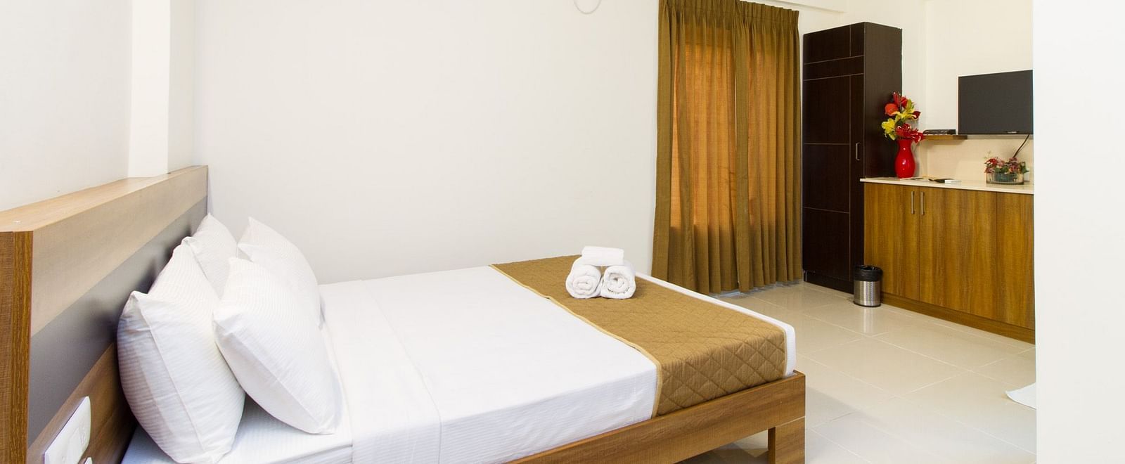 A well-lit hotel room offering a double bed with white and brown linens and a glimpse of a wardrobe and the kitchen area at Sanctum Suites Whitefield, Bangalore.