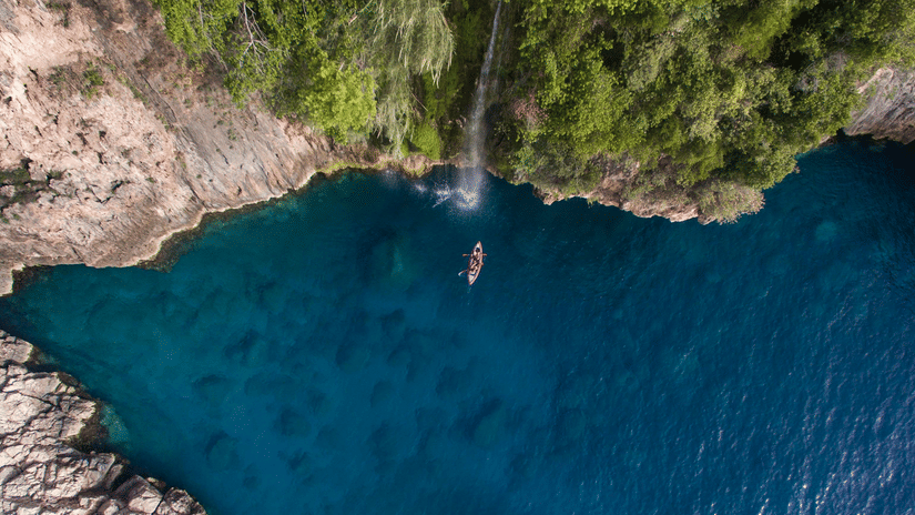 Aerial view of a person kayaking on clear turquoise water in a forested cove with a thin waterfall cascading from green cliffs above.