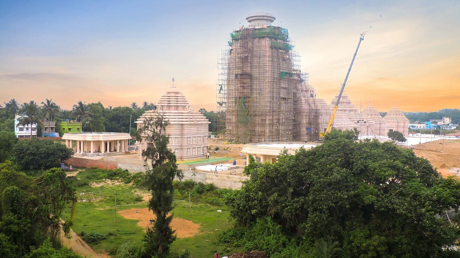 View of Jagannath Temple under construction surrounded by greenery, with scaffolding and cranes in the background, captured during sunset