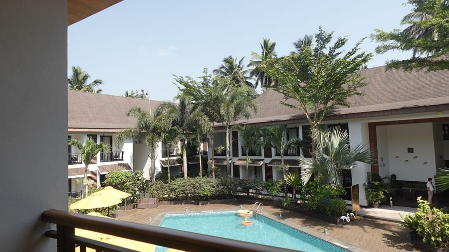 View of a resort courtyard with a swimming pool, surrounded by palm trees and two-story buildings, as seen from a balcony railing - Sobit Sarovar Portico, Goa.