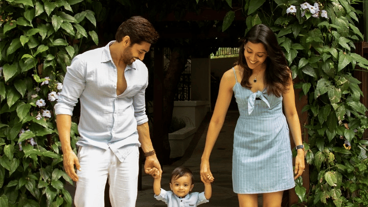 A young family of three walking toward the camera, holding hands. The mother and father smile at their young child, who is between them.