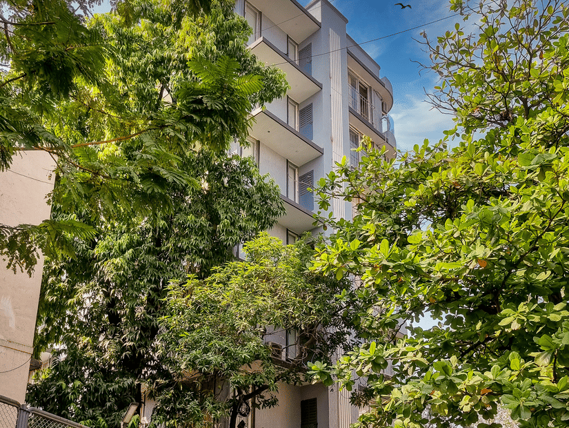 The building exterior of Home2 Residency, Mumbai Airport, with multiple floors, surrounded by trees, tiled walkway in foreground, boundary wall with pattern, and potted plants near entrance.
