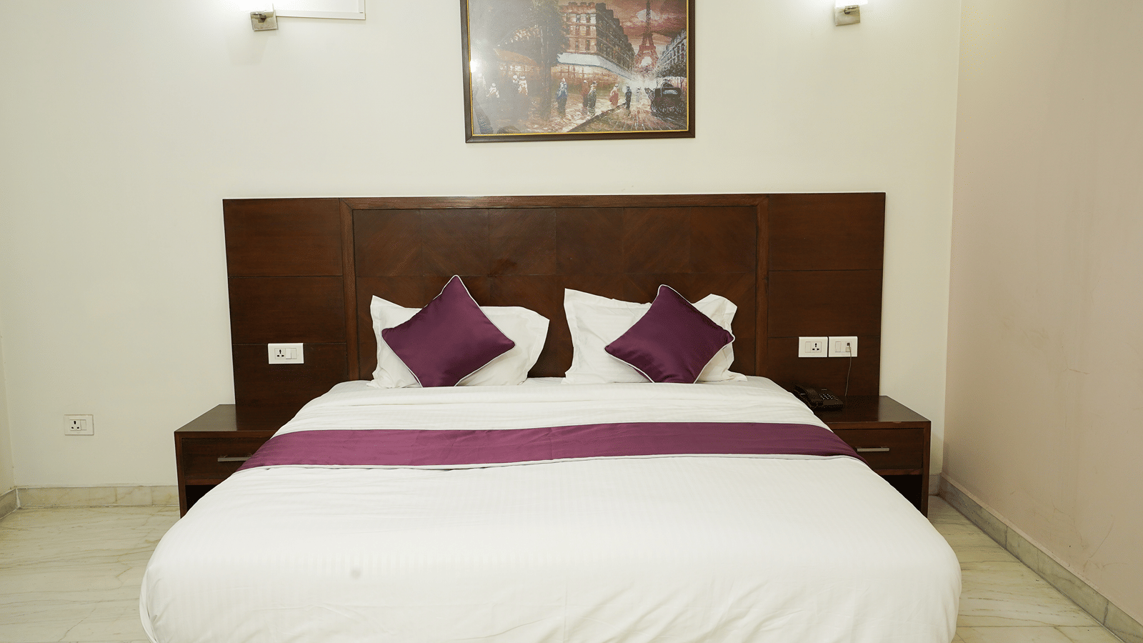 Guest room with a king-size bed, dark wood headboard, purple cushions and runner, wall lamps, and a Paris painting.