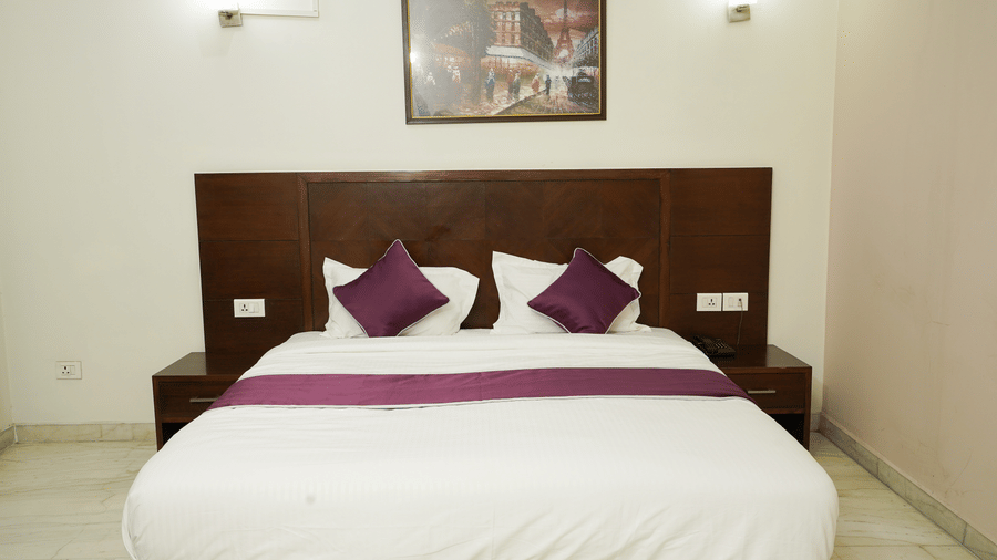 Guest room with a king-size bed, dark wood headboard, purple cushions and runner, wall lamps, and a Paris painting.