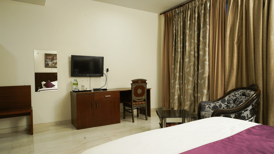 Guest room with a wall-mounted TV on a wood console, a desk with a carved chair, mirror, and a bed with purple runner.