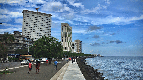 An image of a street of Mumbai near the Marine Drive with high rise buildings where are people are seen walking with a blue sky in the background