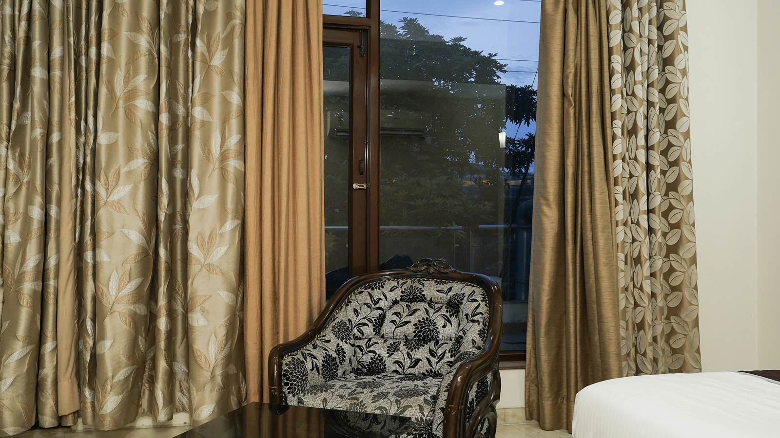 Corner of a guest room with a carved wood floral armchair, a glass table, and a floor-to-ceiling window at dusk.