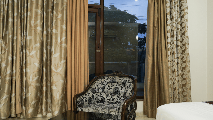 Corner of a guest room with a carved wood floral armchair, a glass table, and a floor-to-ceiling window at dusk.