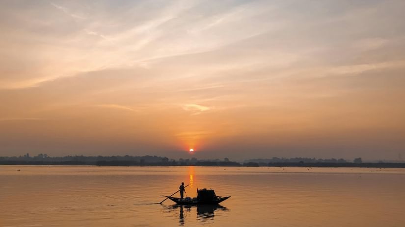 Silhouette of a person along with a boat sailing in a river with the sun in the distance.