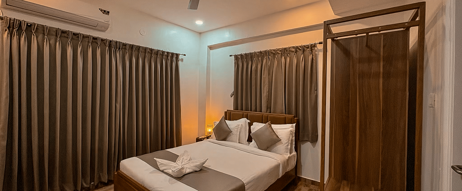 Wide veiw of a bedroom in the Coral 4BHK of Kove Smart Stays featuring wooden flooring, plush bedding, a wardrobe unit, and closed curtains above the headboard of the bed, and one whole wall of the room.