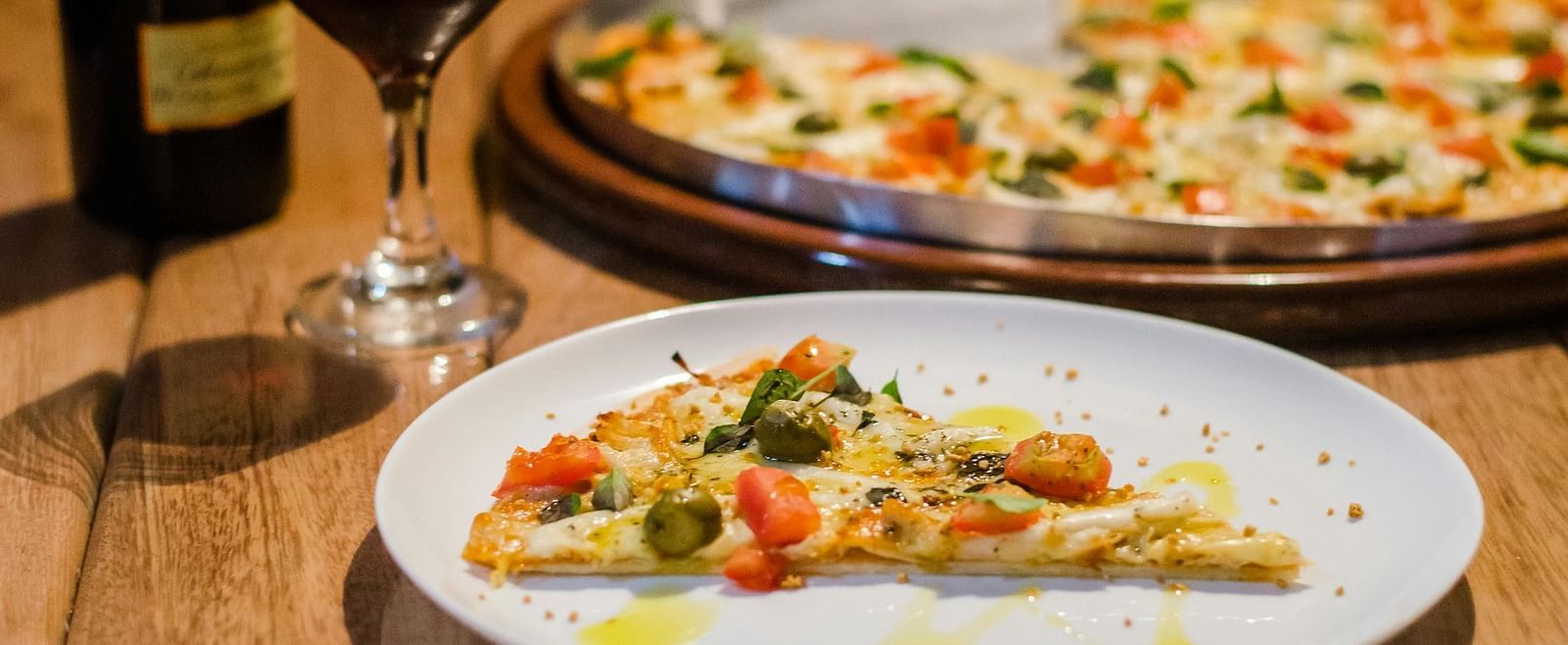 A pizza slice placed next to a wine glass on a wooden table. 