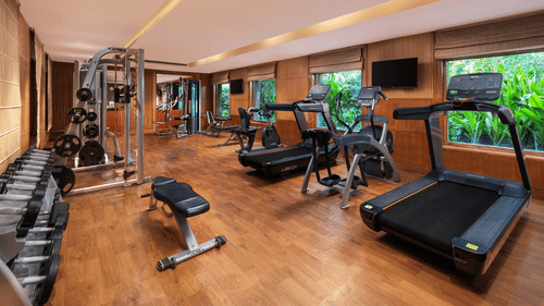 Modern fitness centre with wooden flooring, equipped with treadmills, stationary bikes, weight machines, and a rack of dumbbells, featuring large windows overlooking greenery - Ananta Spa and Resort, Pushkar.