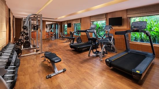 Modern fitness centre with wooden flooring, equipped with treadmills, stationary bikes, weight machines, and a rack of dumbbells, featuring large windows overlooking greenery - Ananta Spa and Resort, Pushkar.