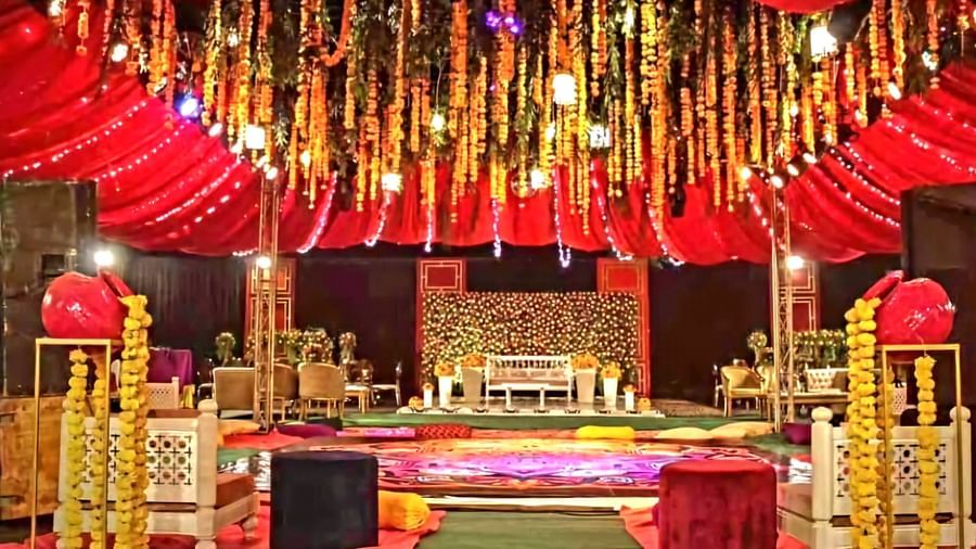An elaborate wedding reception hall is decorated with hanging floral garlands, red and gold drapes, and seating arrangements for guests