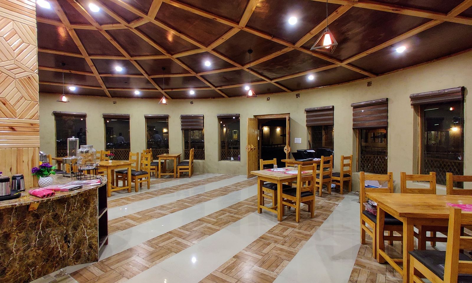The interior of a dining area at White Sands Resort, Ladakh, with wooden tables and chairs, a buffet setup in the background, and decorative lighting fixtures.