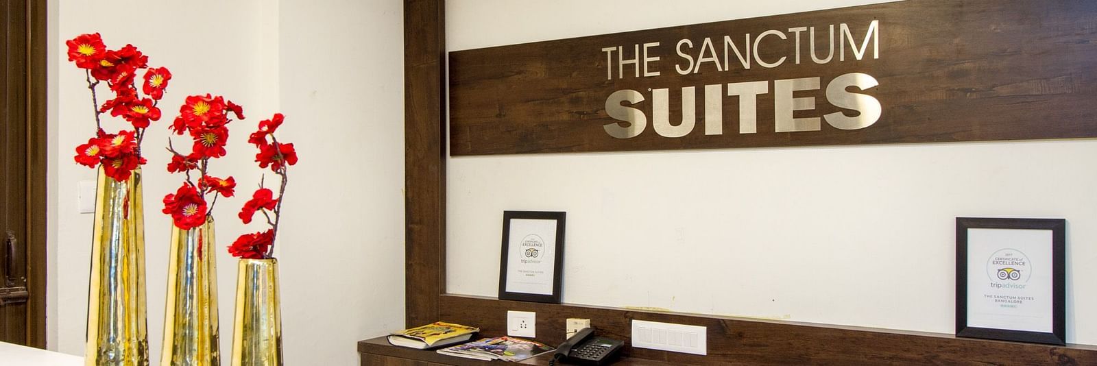 A reception area presenting 'The Sanctum Suites' sign and red flowers placed on a desk at Sanctum Suites Whitefield, Bangalore.