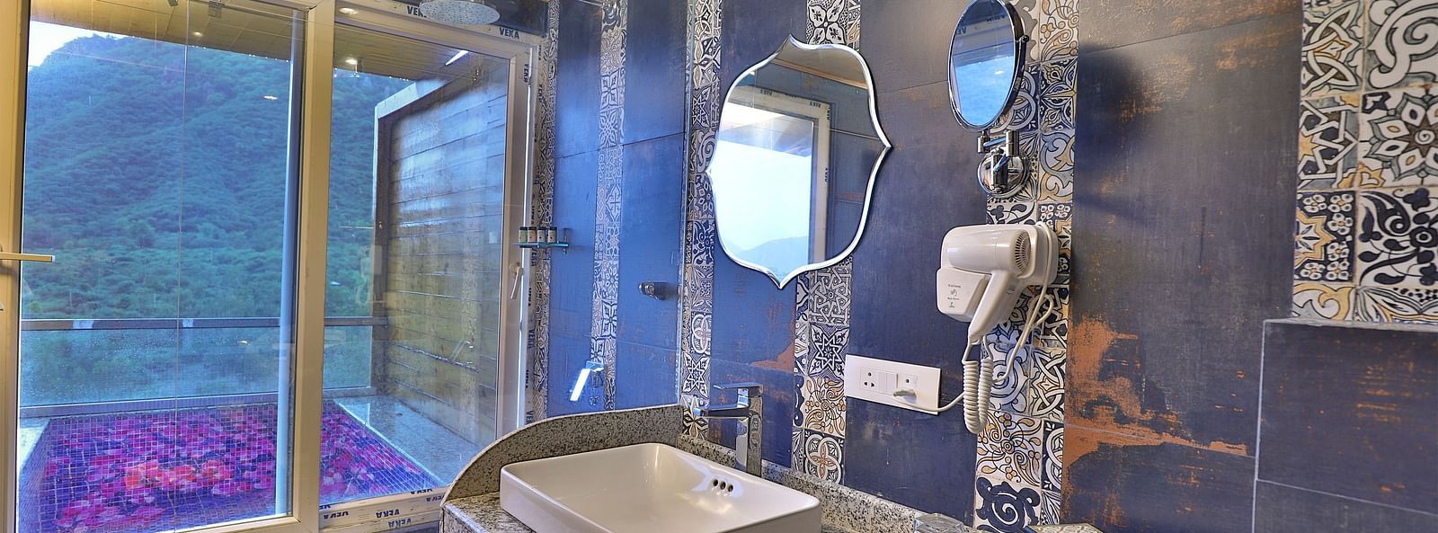 A bathroom with unique tiles, a sink on a speckled counter, and a large window overlooking a lush landscape. - Bamboo Saa Resort & Spa, Udaipur