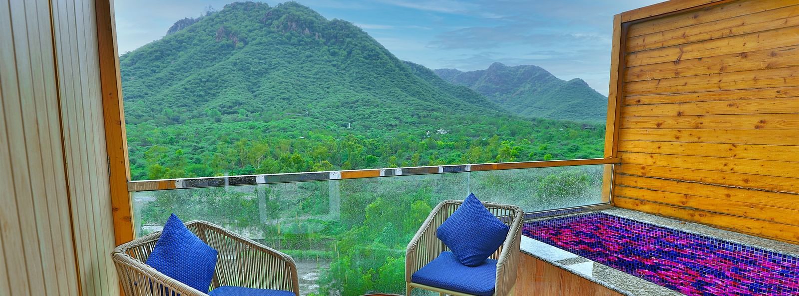 A balcony with two chairs, cushions, and a table overlooks a lush green valley with mountains as a scenery.- Bamboo Saa Resort & Spa, Udaipur
