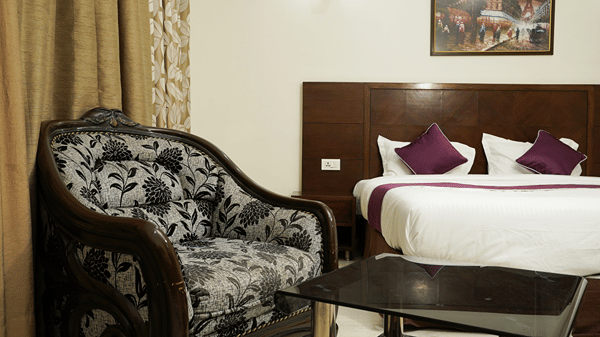 Guest room with a carved wood armchair in floral upholstery, a glass side table, and a bed with purple cushions.