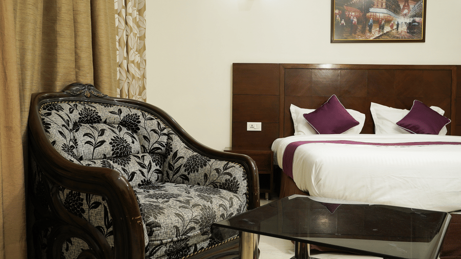 Guest room with a carved wood armchair in floral upholstery, a glass side table, and a bed with purple cushions.