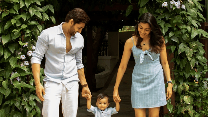A young family of three walking toward the camera, holding hands. The mother and father smile at their young child, who is between them.