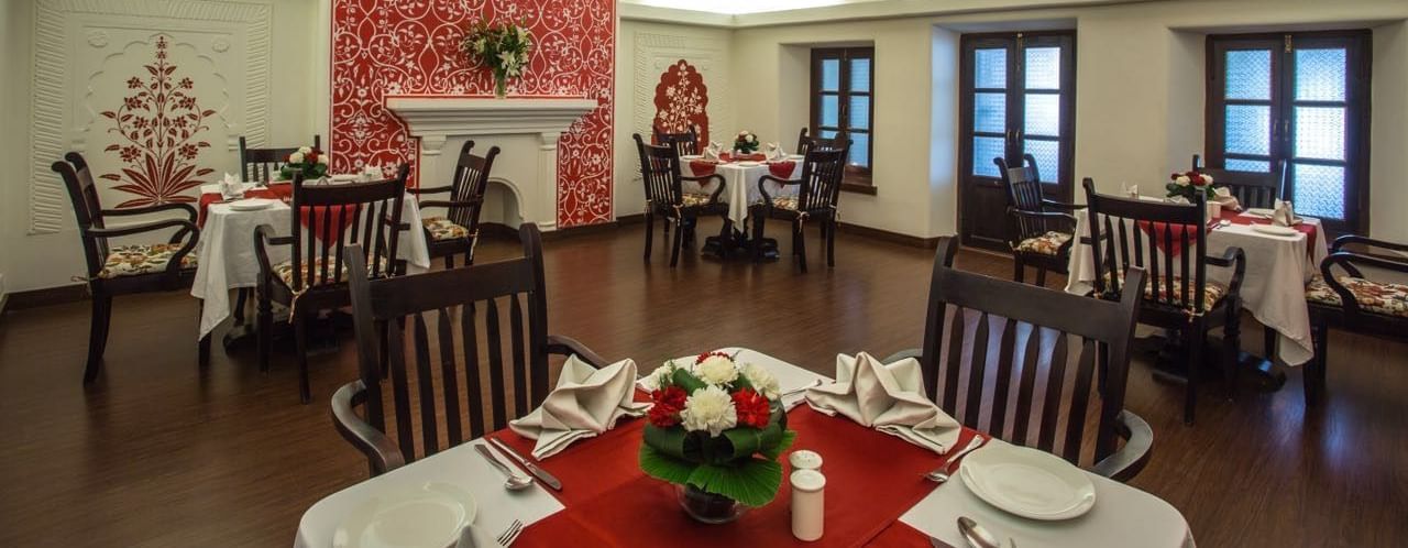An indoor dining hall at Royal Heritage Haveli with high ceilings, several tables with white tablecloths, and a decorative red ceiling.