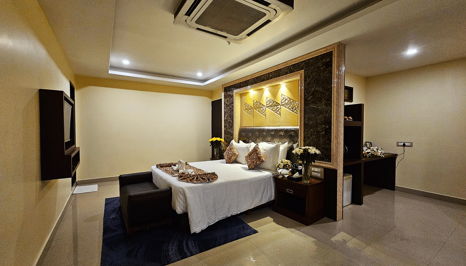 An image of a bedroom with bedside table, a set of table and chairs, flat TV screen, and AC- Sathyam Grand Resort