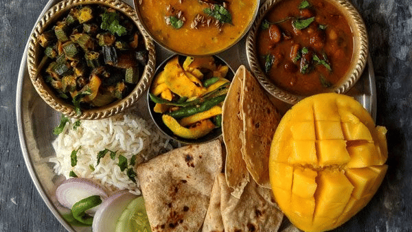 Traditional Indian vegetarian thali featuring dal, bhindi, rajma, rice, rotis, papad, mango, and desserts on a rustic table.