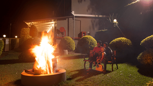 Outdoor bonfire at night. Flames rise from a fire pit, casting light on nearby bushes and a building in the background - Abad Copper Castle, Munnar