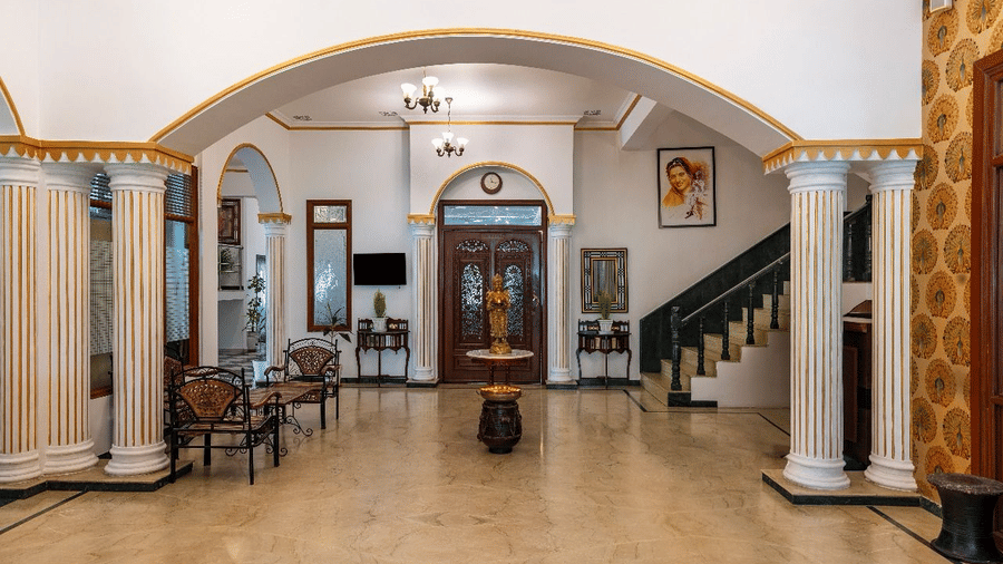 A spacious tiled lobby area with white archways, pillars, seating, and a staircase, at Amantra Shilpi Resort, Udaipur.