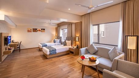 A spacious hotel suite featuring two single beds with blue and white accents, a comfortable seating area with a sofa and armchairs around a small table with fruit, a work desk with a chair, and a television, all on a wooden floor at Lords Inn Motikhavdi