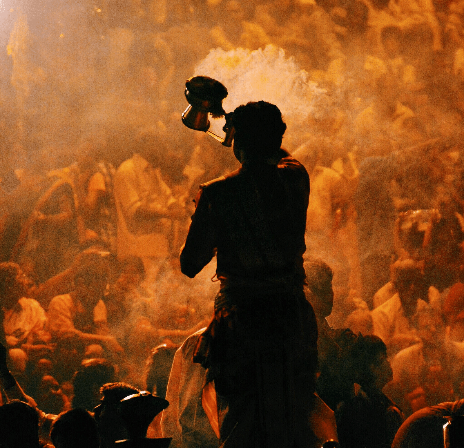 A close-up of a person performing Ganga Aarti, holding a lamp, surrounded by an engaged crowd. The scene captures the spiritual atmosphere