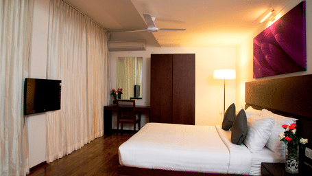 A well lit room featuring a plush bed facing the TV in front of it and a flower vase on the nightstand next to the headboard at Springs Hotel & Spa, Bangalore