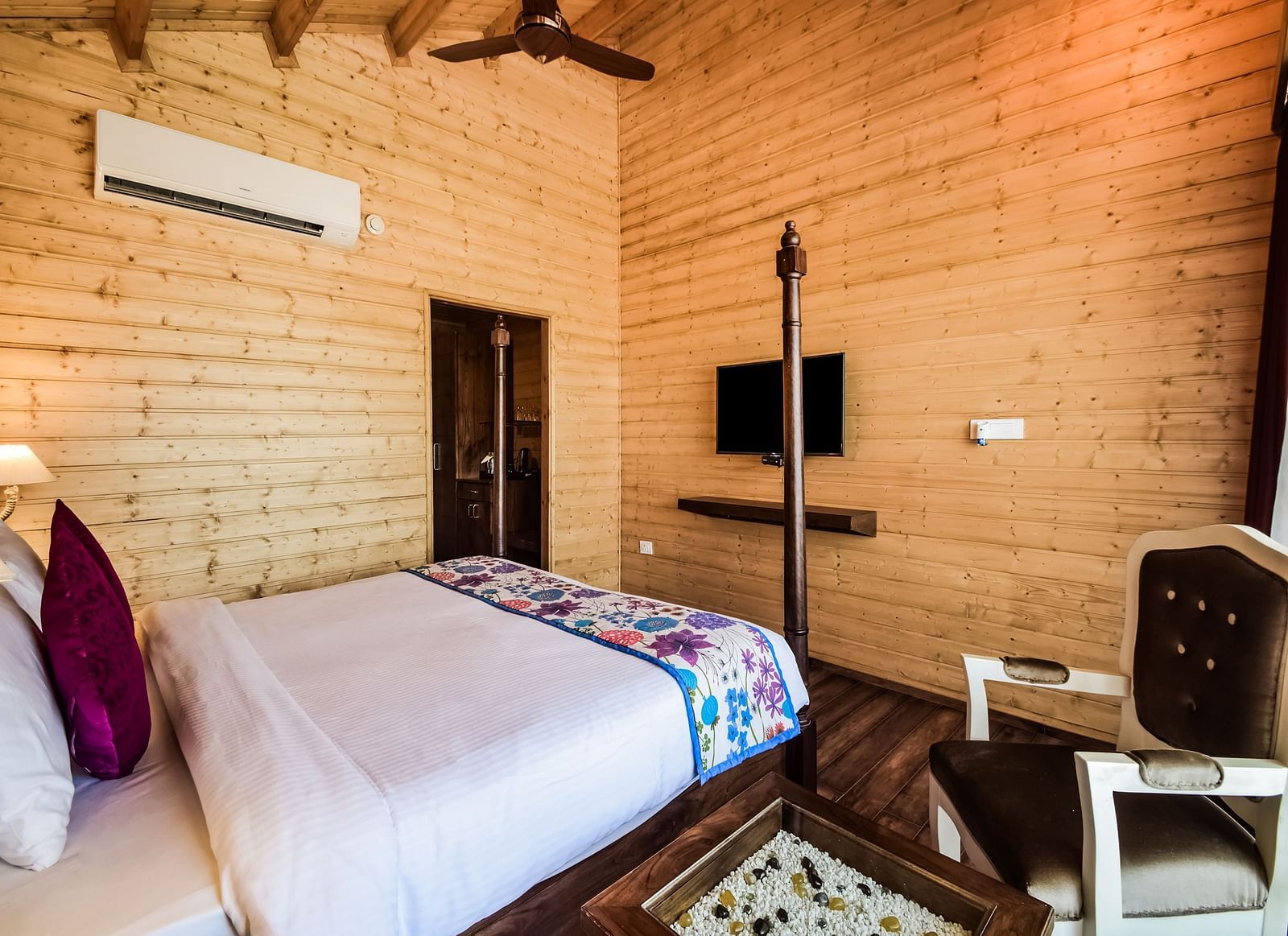 A cosy bedroom with wooden paneling, a plush bed and warm lighting. An air conditioner is mounted on the wall.- Bamboo Saa Resort & Spa, Udaipur
