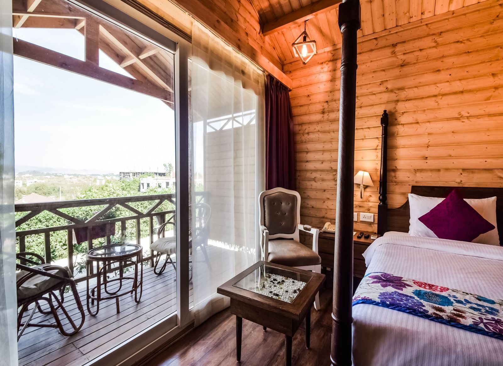 A cosy hotel room with wooden walls, a bed with pillows on it and a balcony overlooking a scenic view.- Bamboo Saa Resort & Spa, Udaipur