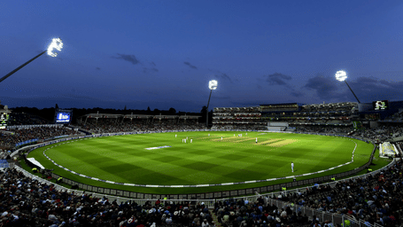Large cricket stadium illuminated by floodlights at night, with a green field and spectators seated around the stands.