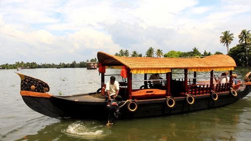 A traditional wooden boat with a curved roof floats on calm water, with palm trees, a cloudy sky, and distant shores in the background.