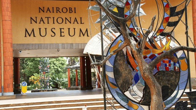 The entrance to the Nairobi National Museum building displays its name in bold letters behind a tall sculpture made of dark metal and decorative circular glass panels.
