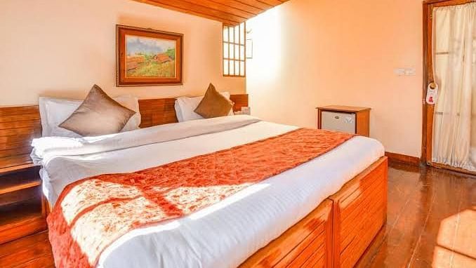 A deluxe room with a large bed adorned with neatly arranged cushions, a bedside table, small windows to the side providing additional brightness, and a photoframe positioned above the bed.