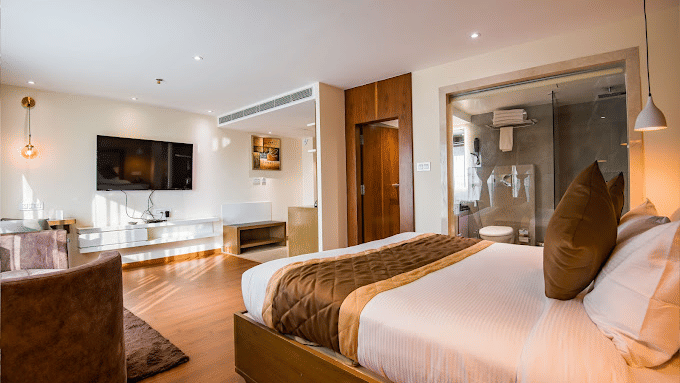 A premium hotel room with a large bed, a wooden table to the side, a wall-mounted TV, and a fancy lamps around the room at Yashshree The Imperial Inn, Siliguri